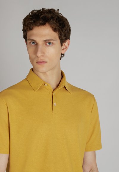 Żółta koszulka polo wykonana z teksturowanej tkaniny z zapięciem na guziki i kołnierzykiem. Model prezentuje neutralne tło i prosty krój.