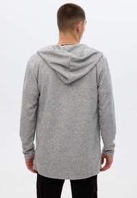 Persona vistiendo un suéter con capucha de manga larga gris, de pie de espaldas a la cámara, combinado con pantalones negros frente a un fondo claro y liso.