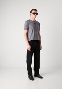 Grå kortærmet t-shirt med et rødt mærkelogo, sorte bukser og sorte solbriller. Minimalistisk design, glat tekstur og tætsiddende silhuet.