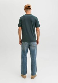 Camiseta de manga corta verde oscuro combinada con jeans holgados de un azul claro. Gorra de color tostado y zapatos marrón claro, destacados por los detalles del material.