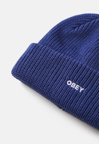 Strikket blå lue med en teksturert design. Har en brettet kant med hvit "OBEY" brodert logo.