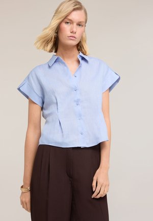 Mujer joven con cabello rubio que lleva una camisa azul clara de manga corta con botones y pantalones anchos oscuros sobre un fondo neutro.