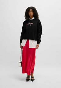 Felpa nera con la scritta "Hugs" in rosa, indossata sopra una camicia bianca a righe, abbinata a una gonna massi rossa in satin e scarpe nere con tacco.