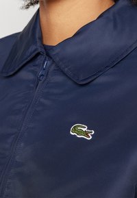 Gros plan d'une personne portant une veste zippée bleu marine avec un logo crocodile vert brodé sur la poitrine.