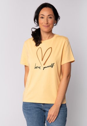 T-shirt en coton de couleur jaune doux avec des manches courtes, affichant un cœur marron et un texte noir disant "aime-toi." Coupe décontractée et texture lisse.