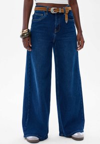 Szerokie niebieskie dżinsy z wysokim stanem, z brązowym paskiem i srebrną klamrą. Gładka faktura z przednimi kieszeniami.