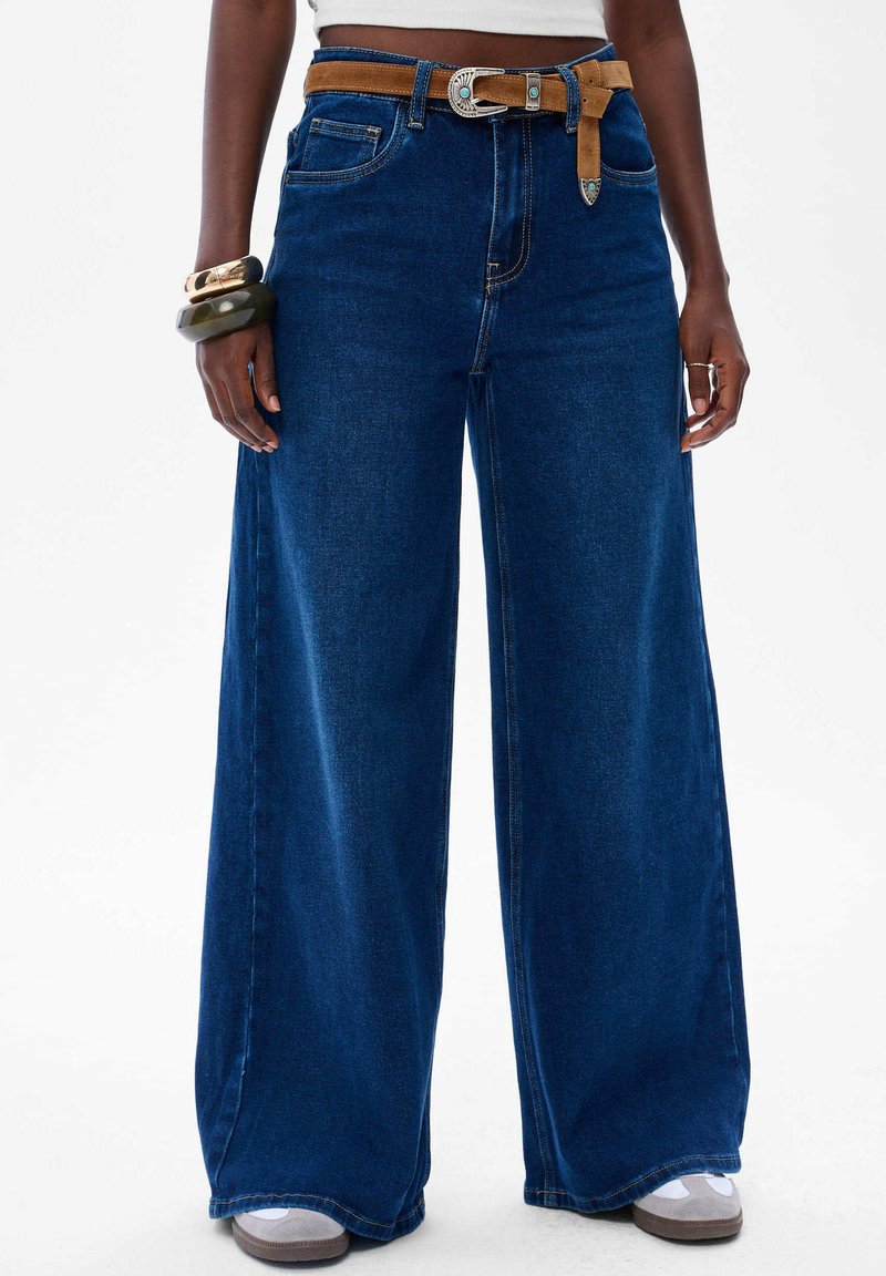 Szerokie niebieskie dżinsy z wysokim stanem, z brązowym paskiem i srebrną klamrą. Gładka faktura z przednimi kieszeniami.