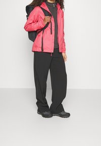 Chaqueta impermeable rosa con capucha, combinada con una camisa oscura de manga larga y pantalones negros holgados. Zapatos de senderismo negros y una mochila completan el conjunto.