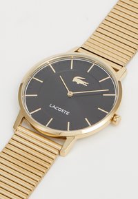 Auksinis laikrodis su juodu ciferblatu, turintis aukso valandų žymeklius, Lacoste logotipą ir auksinį tinklinį dirželį. Apvalus korpusas su skaidriu stiklu.