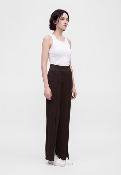 Femme aux cheveux courts et foncés portant un débardeur blanc, un pantalon noir large taille haute et des baskets blanches, debout devant un fond uni.