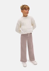 Suéter blanco de punto trenzado con un cuello alto y mangas largas, combinado con pantalones de pierna ancha de color rosa claro y zapatillas blancas.
