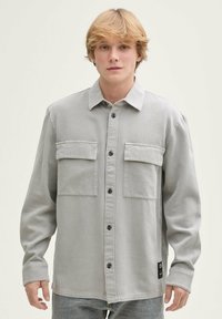 Hellgraues Hemd mit Knöpfen aus strukturiertem Stoff, ausgestattet mit zwei Brusttaschen, schwarzen Knöpfen und einem Detail mit einem seitlichen Etikett.