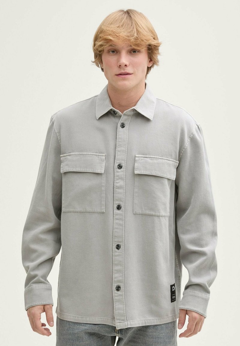 Hellgraues Hemd mit Knöpfen aus strukturiertem Stoff, ausgestattet mit zwei Brusttaschen, schwarzen Knöpfen und einem Detail mit einem seitlichen Etikett.