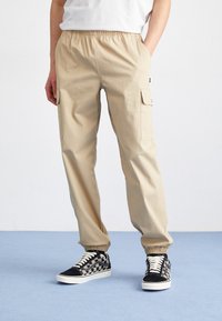 Khaki Cargo-Hosen mit elastischem Bund, Seitentaschen und schmal zulaufenden Beinen, kombiniert mit schwarz-weißen gemusterten Sneakern auf einem blauen Boden.