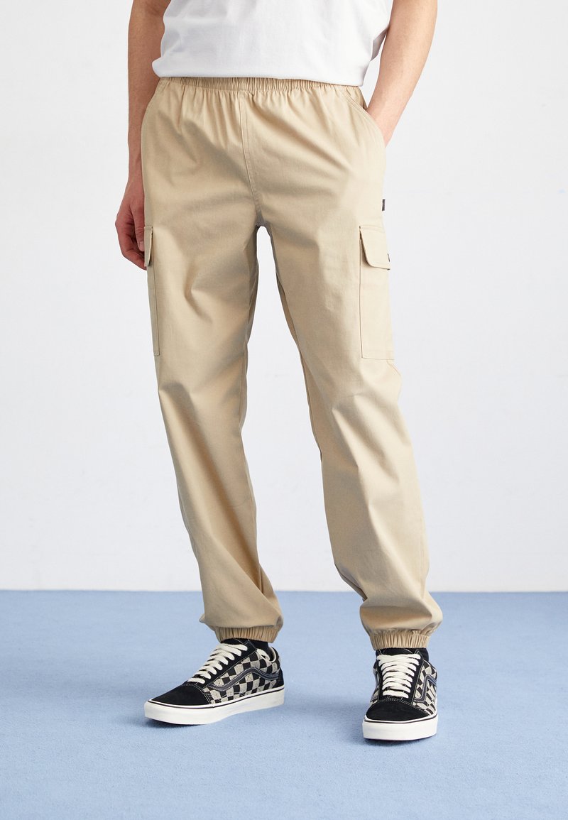 Khaki Cargo-Hosen mit elastischem Bund, Seitentaschen und schmal zulaufenden Beinen, kombiniert mit schwarz-weißen gemusterten Sneakern auf einem blauen Boden.