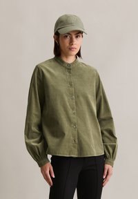 Chemise en velours côtelé vert olive avec un col montant, fermeture à boutons et manches froncées. Associée à une casquette assortie avec un détail brodé.