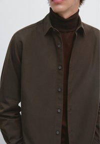 Chemise marron à boutons fabriquée dans un tissu lisse, avec un col et des boutons foncés, superposée à un col roulé marron. Design minimaliste.
