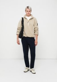 Beige fleece genser med halv glidelås og ribbestrikkede mansjetter, kombinert med marineblå joggebukser og hvite sneakers med svarte detaljer.
