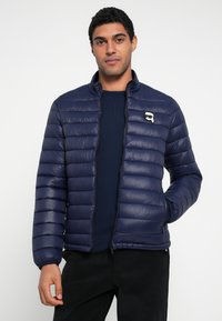 Granatowa kurtka puchowa z poziomymi przeszyciami, zamkiem błyskawicznym, wysokim kołnierzem i detalem z logo na piersi. Gładka, błyszcząca tekstura i dopasowany krój.