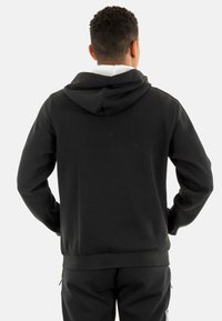 Felpa nera con cappuccio dal tessuto morbido, dotata di tasca a marsupio e polsini a coste. La vista posteriore mostra un design aderente e un cappuccio regolabile.