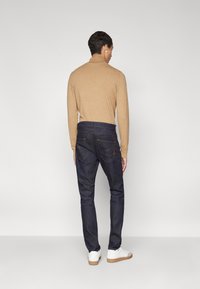 Jeans de mezclilla azul oscuro con un corte recto, combinados con una camisa de manga larga marrón claro. Presenta bolsillos y detalles de costura sutiles. Zapatillas blancas.