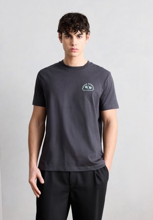 Mladý muž s kučeravými vlasmi, oblečený v tmavosivej tričku s logom "Mood Sports" a čiernych nohaviciach, stojaci na jednoduchom svetlosivom pozadí.