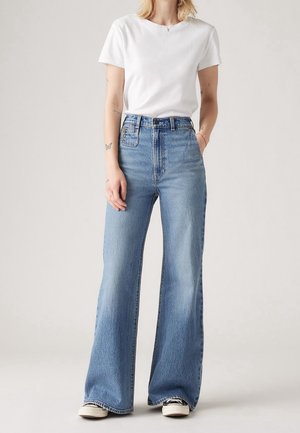 Personne portant un jean bleu taille haute à jambes larges, un t-shirt blanc à manches courtes et des baskets, debout devant un fond clair uni.