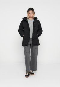 Chaqueta acolchada negra con capucha, con un diseño de acolchado y botones a presión. Llevada sobre un suéter gris claro y pantalones cargo grises.