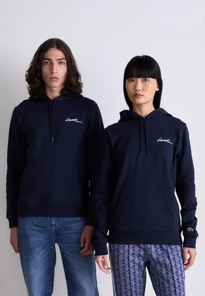 Marina blå huvtröjor i bomullsblandning, med en framficka och en logotyp på bröstet. En modell har på sig jeans, den andra mönstrade byxor.