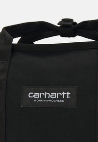 Czarna torba z mocnym designem. Zawiera wyraźną czarną metkę z białym tekstem: "carhartt WORK IN PROGRESS." Regulowany pasek u góry.