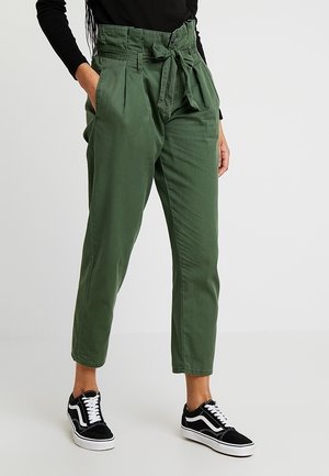 Osoba nosząca oliwkowe zielone spodnie z wysokim stanem, wiązane z przodu i plisami, w połączeniu z czarno-białymi tenisówkami oraz czarną bluzką.