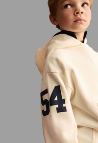 Krämfärgad hoodie med ett svart nummer "54" på vänster ärm. Har en dragsko för huvan och ett mjukt, texturerat tyg.