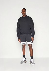 Uomo in piedi, che indossa una felpa nera a maniche lunghe, pantaloni corti sportivi neri con strisce bianche, calze nere e scarpe da ginnastica bianche, su sfondo neutro.