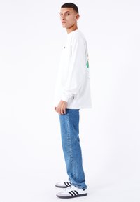 Hvid langermet T-shirt med et farverigt grafik på ryggen, parret med blå denim jeans og hvide sneakers med sorte striber.