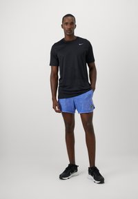 Schwarzes Nike-Shirt mit kurzen Ärmeln, blaue gemusterte Shorts und schwarze Turnschuhe. Fokus auf sportlichem Design und bequemem Schnitt.