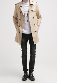 Béžový trenčkot s dvouřadými knoflíky, ramenními klapkami a bočními kapsami, nošený přes bílé tričko s potiskem a černé džíny.