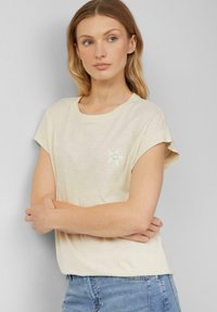 Beige t-shirt met korte mouwen en een ronde halslijn, met een kleine groene ster borduursel op de borst. Zachte, gestructureerde stof.