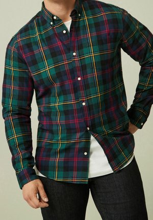 Homme portant une chemise à carreaux verte, bleu marine, rouge et jaune, boutonnée, par-dessus un t-shirt blanc avec un pantalon foncé, main reposant près de la taille.