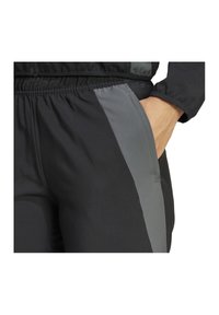Schwarze Sporthose mit elastischem Bund und Seitentaschen, mit einem grauen Streifen, der entlang des äußeren Oberschenkels verläuft. Glatter, dehnbarer Stoff.