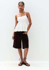 Hvid ærmeløs top med lagdelt design, parret med mørk bordeaux knælange shorts og sorte flade sandaler med vævede detaljer.