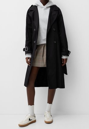 Person wearing a black trench coat over a gray hoodie, plaid pleated mini skirt, white socks, and white sneakers with gum soles.