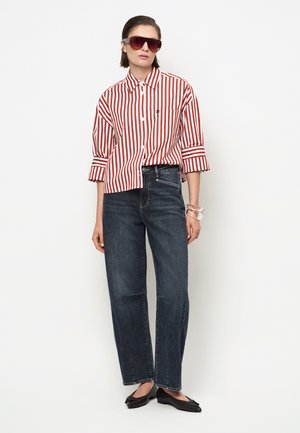 Vrouw draagt een rood-wit gestreept shirt, donkere high-waist jeans, zwarte platte schoenen, oversized zonnebril en een doorzichtige armband om haar pols.