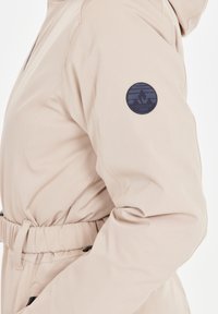 Beige jacka med texturerad yta, med insydd midja, huva och en mörkblå cirkulär logotyp på vänster ärm.