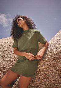 Robe t-shirt vert olive à manches courtes, coupe décontractée et col rond, portée avec un collier de perles et des lunettes de soleil rondes, sur un fond rocheux.