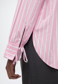 Κοντινή λήψη ενός χεριού δίπλα σε μανσέτα πουκάμισου ροζ και λευκών ριγών με δεμένους κορδέλες, σε συνδυασμό με μαύρο παντελόνι.