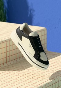 Sort og kremfarget skinnsneaker med rund tå, perforerte detaljer, flate snøringer og en teksturert såle, vist på en rutete overflate.