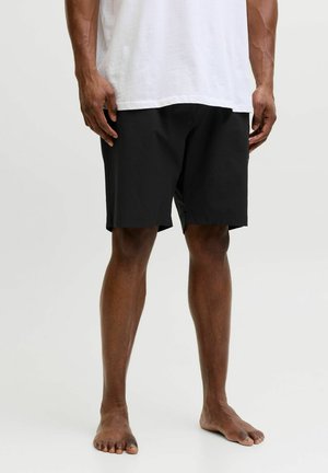 Hombre vestido con pantalones cortos negros hasta la rodilla y una camisa blanca, de pie descalzo sobre un fondo claro y liso.