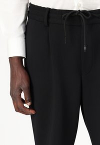 Black patterned trousers with an elastic waistband, drawstring closure, and side pockets, paired with a white shirt sleeve.