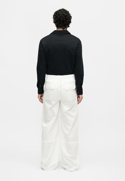 Άτομο με σγουρά μαλλιά που φορά μαύρο μακρυμάνικο πουκάμισο και φαρδιά λευκά παντελόνια, όρθιο με την πλάτη προς ένα απλό φόντο.