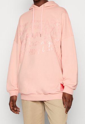Personne portant un sweat à capuche rose clair avec un motif de texte discret et un pantalon beige, debout devant un fond clair uni.